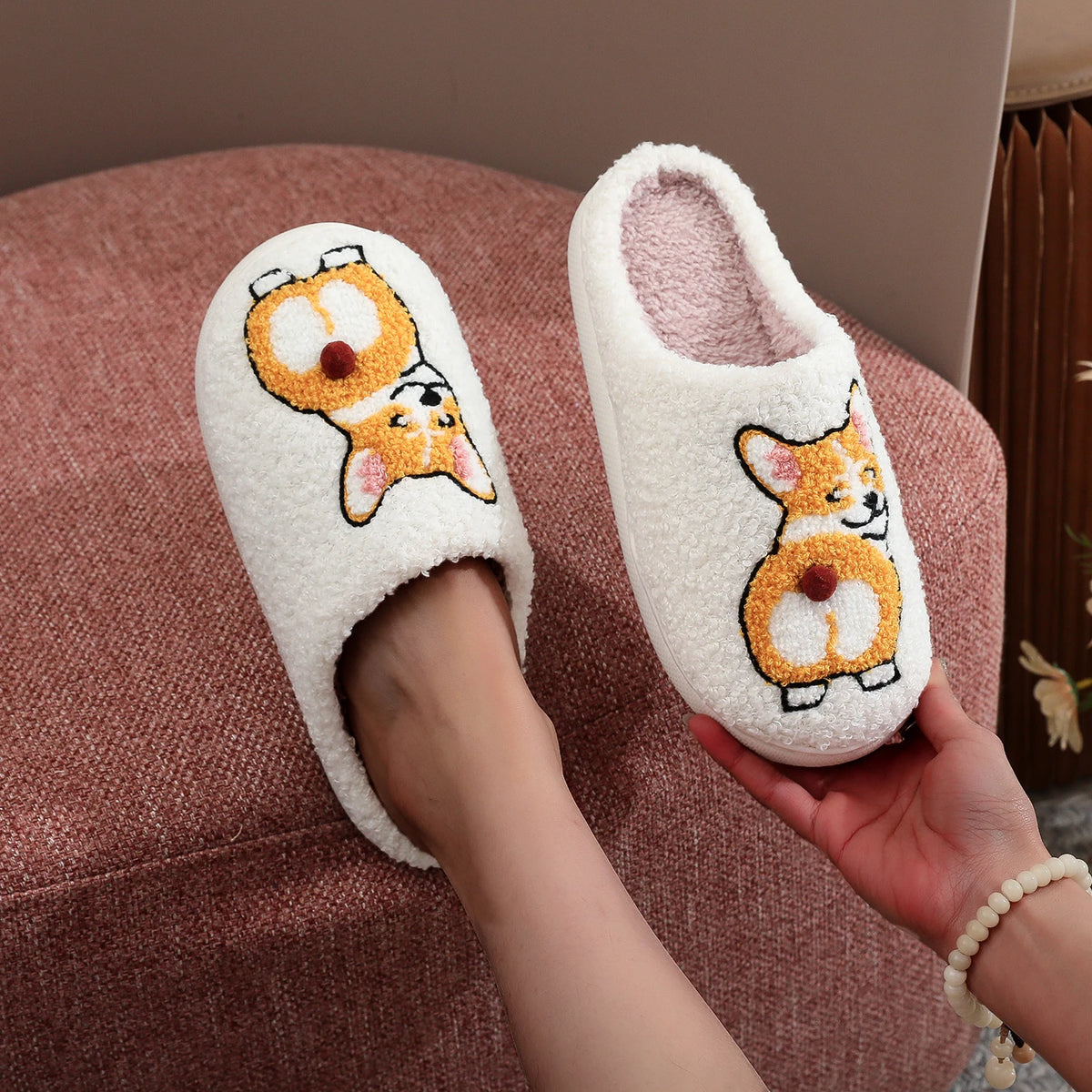 A pair of fluffy white slip-on house slippers with embroidered corgi designs, shown with one worn on a foot and the other held by a hand against a pink upholstered chair background.