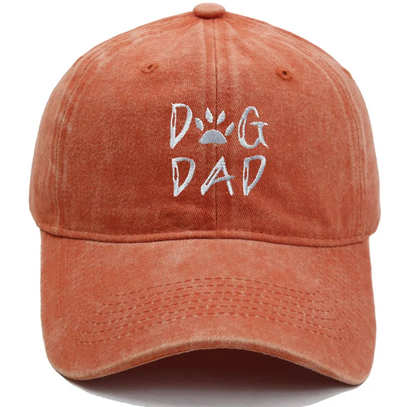 A washed terracotta orange baseball cap featuring 'DOG DAD' embroidered on the front in white, with a paw print graphic inside the letter O.