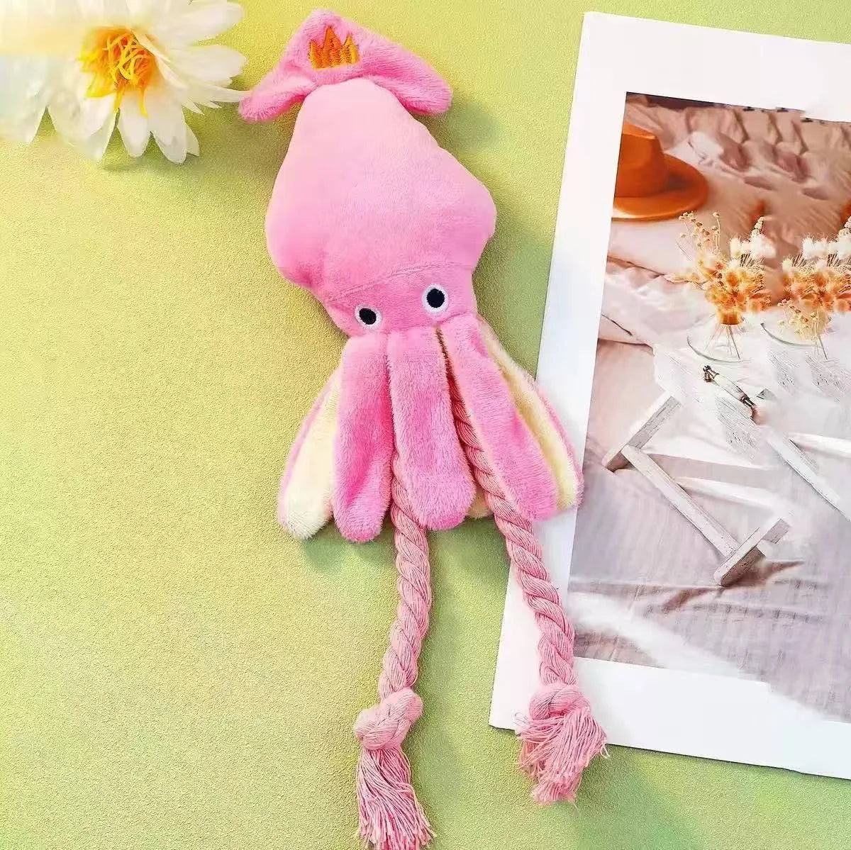 A pink plush octopus dog toy with long rope tentacles and a small orange ring on the head, styled on a light green surface next to a white flower and a printed photo card.