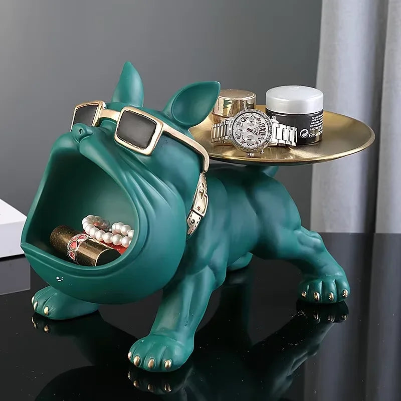 A teal dog-shaped jewelry organizer and tray holder, styled with gold-tone sunglasses, a gold tray on its back, and various jewelry and watch accessories inside and on top, set against a dark gray background.